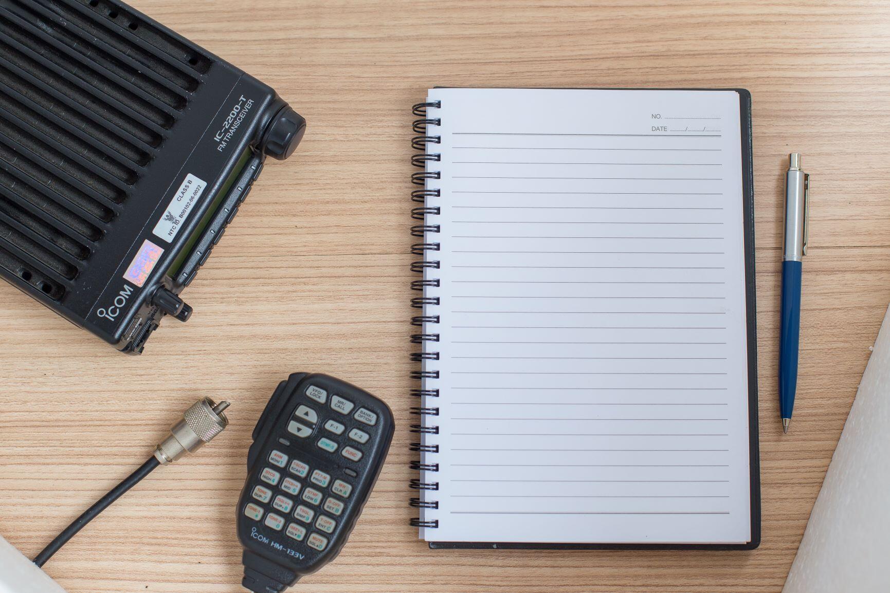 An Icom Radio sitting on a wooden desk with a pen and paper sitting next to the microphone.